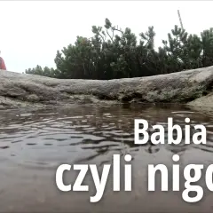 Mglisty dzień na Babiej Górze. Dwie osoby w kurtkach przeciwdeszczowych stoją za kamieniem, obok kałuży.