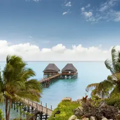 Dwa domki na wodzie połączone pomostem, otoczone palmami. Spokojne morze i błękitne niebo z chmurami. Idealne miejsce na relaks, nawet podczas pory deszczowej na Zanzibarze.