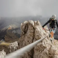 Kobieta w kasku i uprzęży pokonuje skalną grań, zabezpieczona liną. To właśnie via ferrata – bezpieczna droga wspinaczkowa.