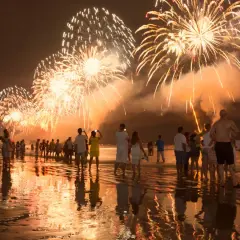 Ludzie na plaży podziwiają spektakularny pokaz fajerwerków. To idealny sylwester w ciepłych krajach, z odbiciami świateł w wodzie.