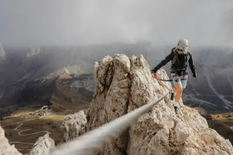 Kobieta w kasku i uprzęży pokonuje skalną grań, zabezpieczona liną. To właśnie via ferrata – bezpieczna droga wspinaczkowa.