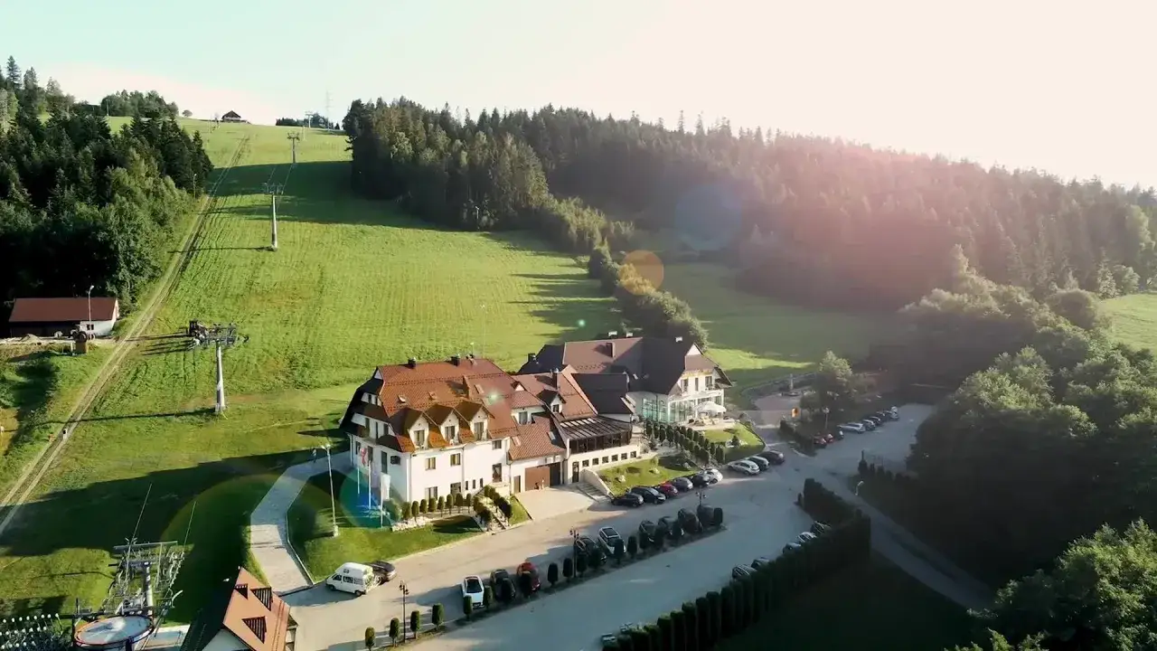 Hotel ze stokiem narciarskim, otoczony zielonymi wzgórzami i lasem. Widoczna kolejka linowa i parking pełen samochodów.